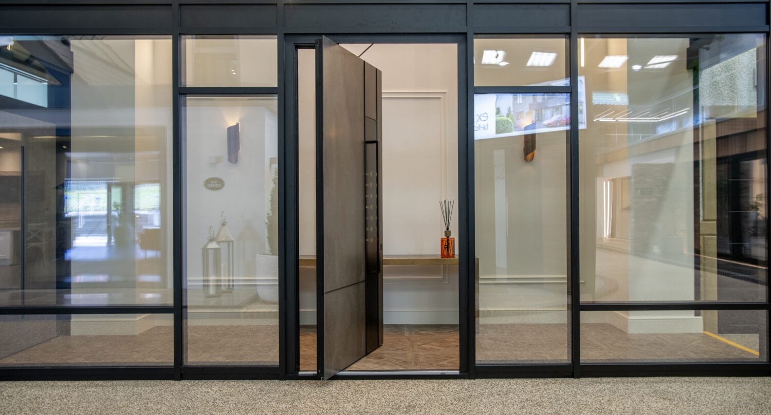 Glass wall with a large modern pivot front door, partially open, revealing a minimal interior with a wooden console table and orange decorative sticks. The speckled floor and bright lighting enhance these bespoke glass solutions.