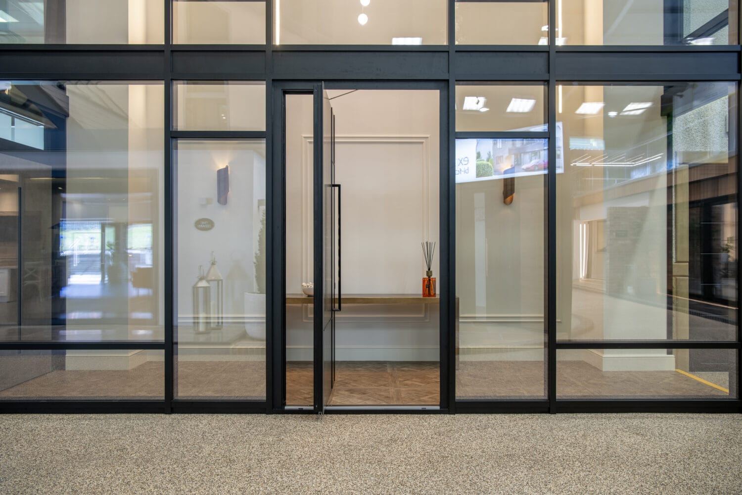 A modern glass-walled office with a glass door, black metal frames, and a wooden table inside. The table holds a decorative vase with diffuser sticks and a bowl. The room features bright lighting, glass roofing, and a speckled floor.