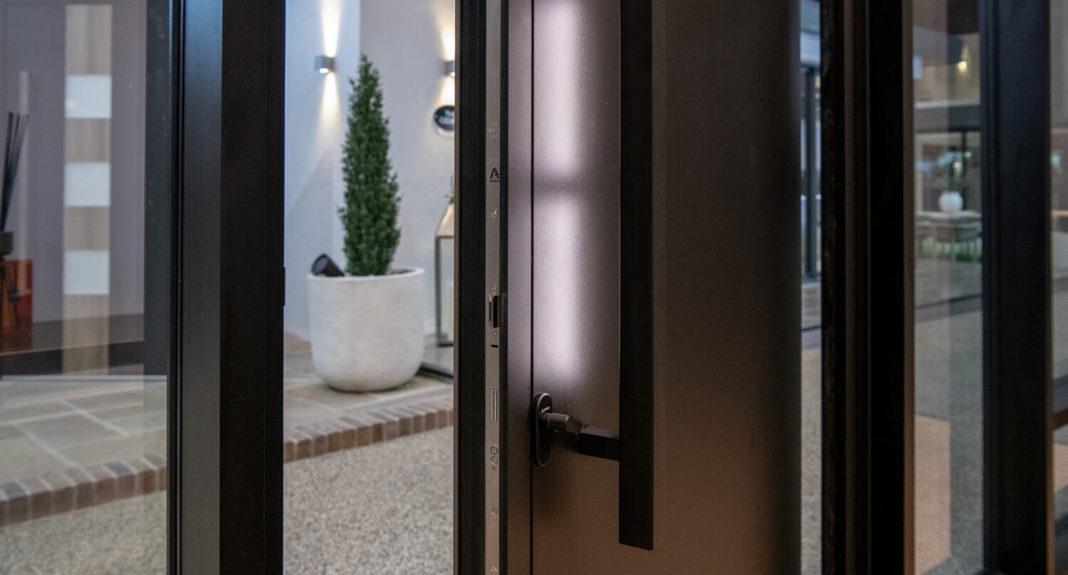 A close-up of a modern black bifold door slightly ajar, revealing a patio with a white planter holding a tall green plant, wall lights, and contemporary decor in the background.