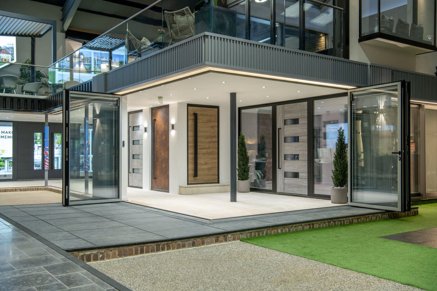 Modern showroom displaying contemporary glass doors, bifold doors, and large windows, with sleek lines, potted plants, and a mix of tiled and artificial grass flooring for a clean, open, and stylish architectural environment.