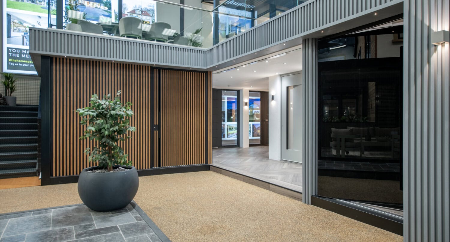 Modern indoor showroom with large windows, wooden paneling, a potted plant, tiled floor, sliding doors, and glass balcony railing. Light from digital displays and recessed lighting creates a bright, contemporary atmosphere.