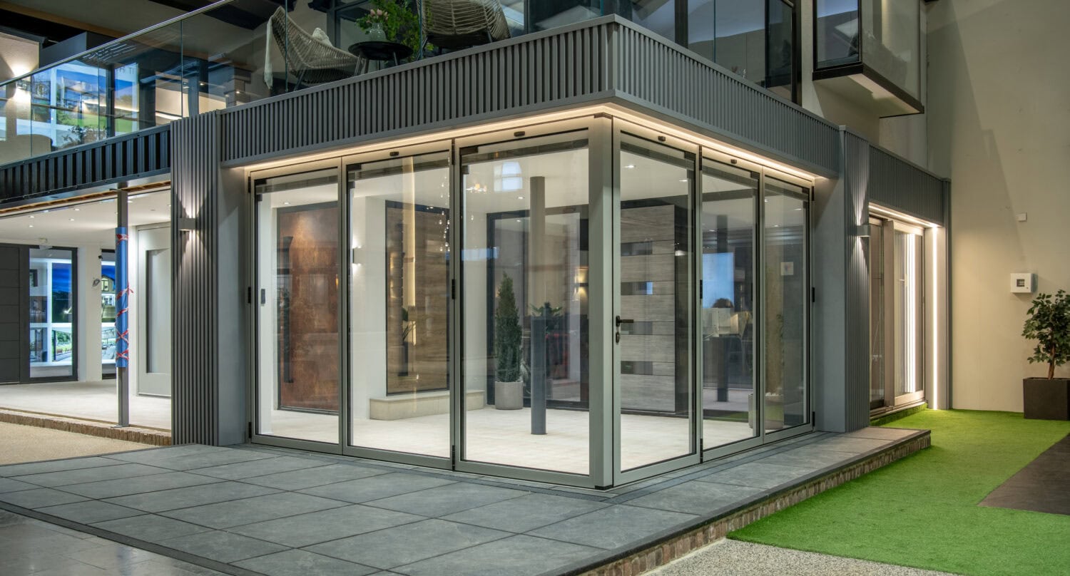 Modern building with large bifold doors and windows, featuring a balcony with outdoor chairs above and a mix of tile and artificial grass flooring outside. The well-lit interior includes potted plants for a fresh touch.