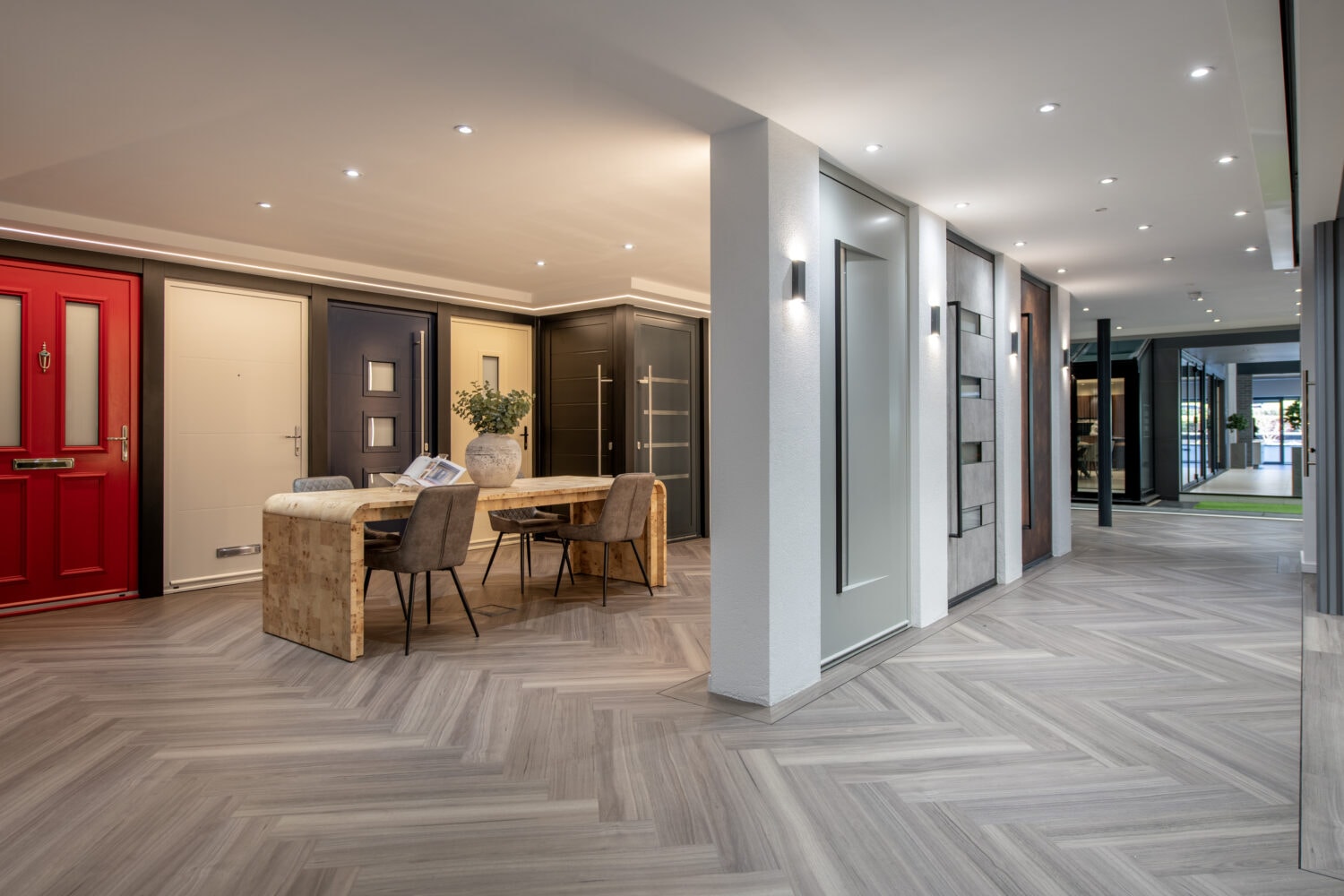A modern showroom displays various front doors and sliding doors, with a table and chairs arranged in the center on a herringbone-patterned floor, under bright ceiling lights.