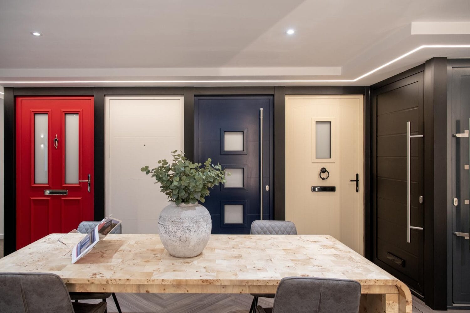 A modern showroom features a table with a large vase of greenery and four chairs, set in front of a display of stylish bifold doors in red, white, navy blue, and cream beneath elegant glass roofing.