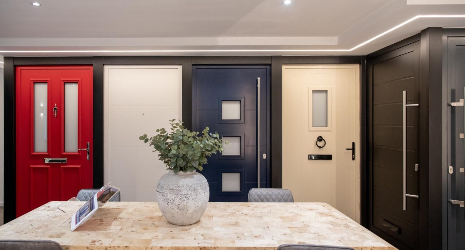 A modern showroom features a table with a large vase of greenery and four chairs, set in front of a display of stylish bifold doors in red, white, navy blue, and cream beneath elegant glass roofing.