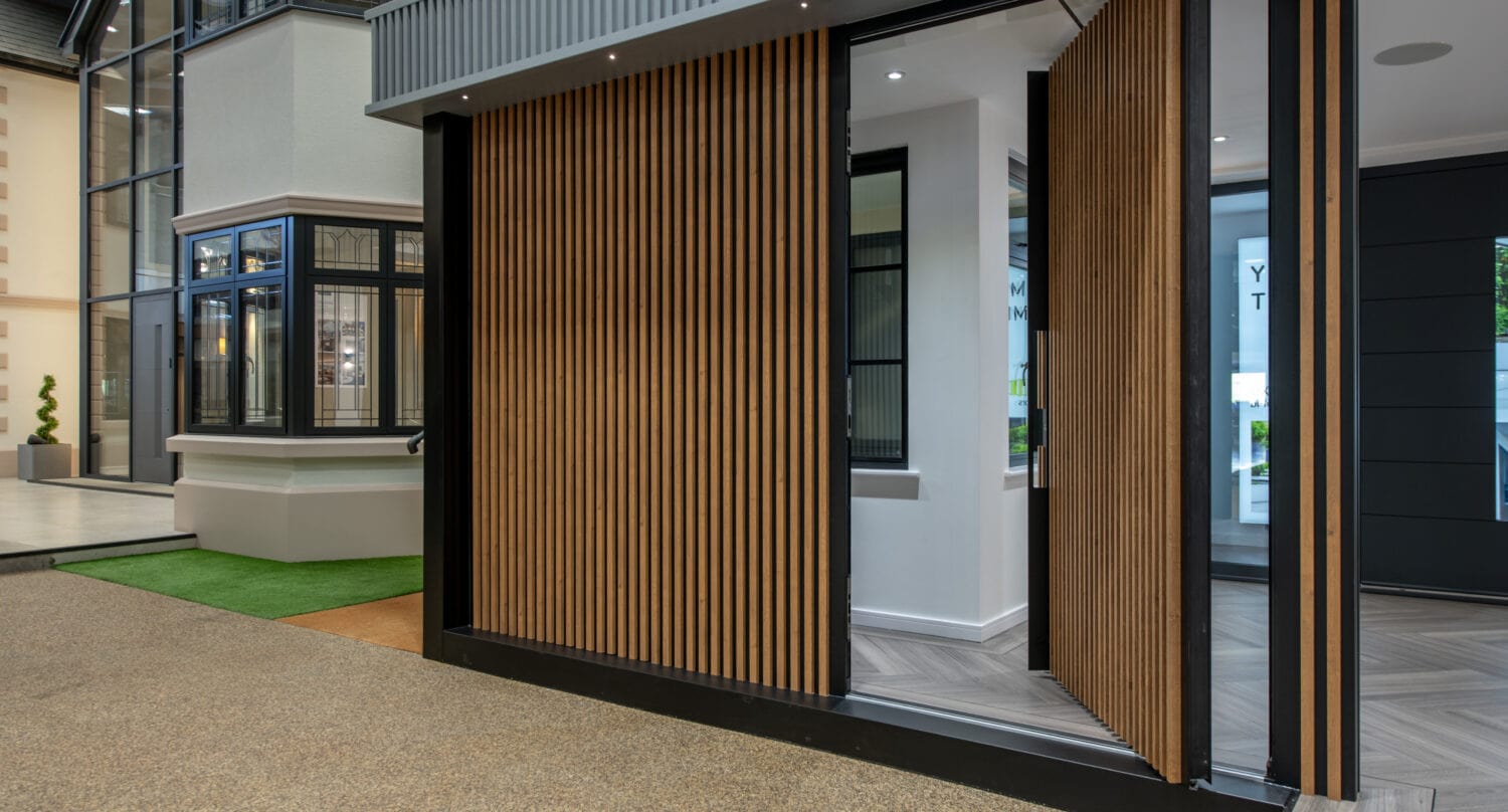 Modern building entrance with vertical wooden slats forming a pivoting door, partially open, revealing a stylish interior with large windows and sliding doors, plus a mix of textured flooring and greenery outside.