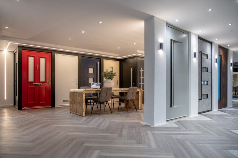 A modern showroom displays various stylish front doors and sleek sliding doors, including a prominent red door. A wooden table with chairs and a plant sits in the center, beneath recessed ceiling lights on a herringbone-patterned floor.