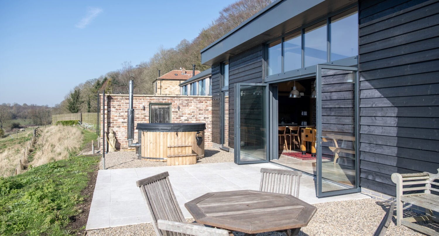 A modern black-clad building with large bifold doors opens onto a patio with a wooden table and chairs, a hot tub, and countryside views on a sunny day.