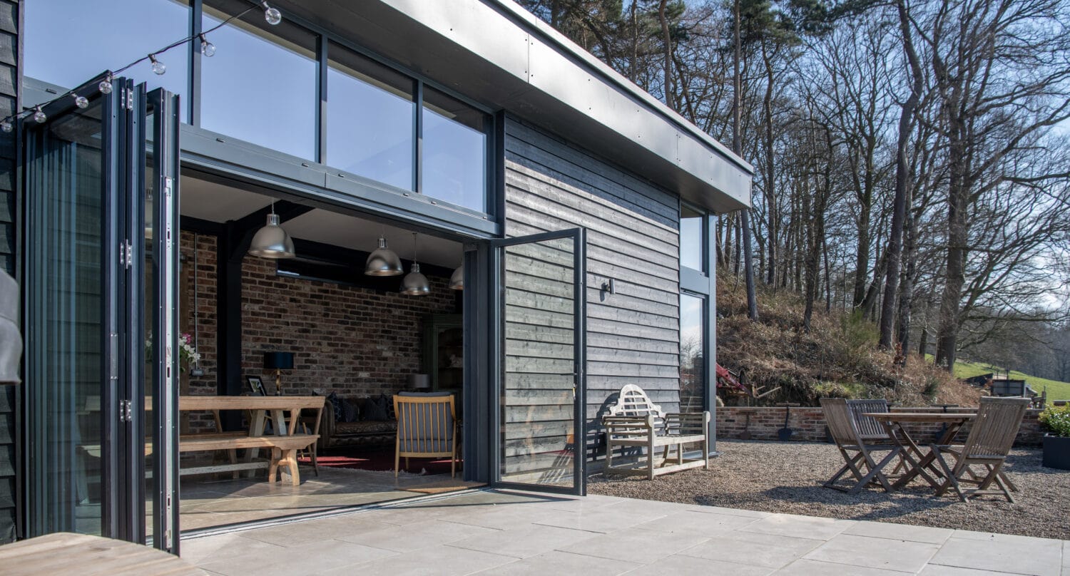 Modern house with large bifold doors open to a spacious patio, featuring outdoor seating and dining areas. The setting is surrounded by trees and greenery under a clear, sunny sky.
