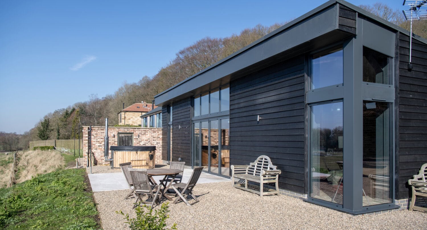A modern black wooden house with large glass windows and sliding doors, outdoor seating, and a hot tub on a gravel patio, surrounded by greenery and trees under a clear blue sky.