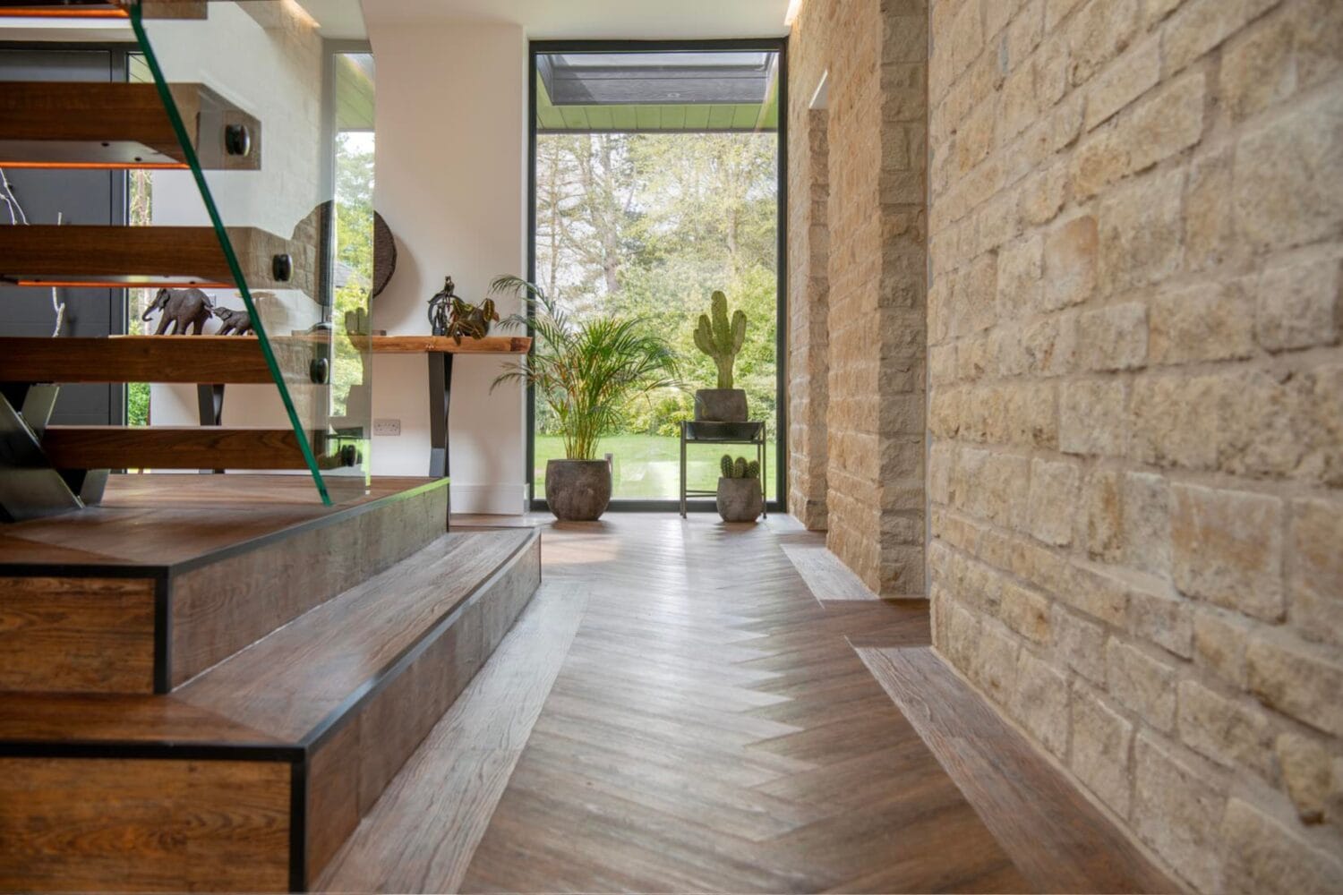 Modern entryway with wood and glass staircase, stone wall, herringbone wood floor, potted plants, and large windows overlooking a green garden. Sliding doors allow natural light to fill the space, highlighting the contemporary design.