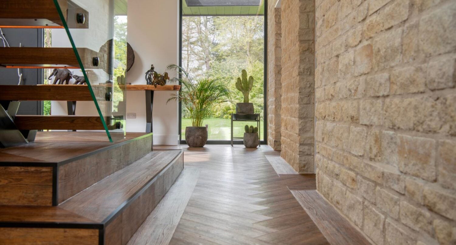 Modern entryway with wood and glass staircase, stone wall, herringbone wood floor, potted plants, and large windows overlooking a green garden. Sliding doors allow natural light to fill the space, highlighting the contemporary design.