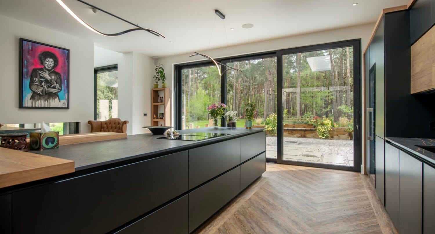 Modern kitchen with sleek black cabinets, wood accents, and herringbone flooring. Sliding doors open to a garden patio, and a colorful portrait hangs on the wall. Natural light from bespoke glass solutions fills the space.
