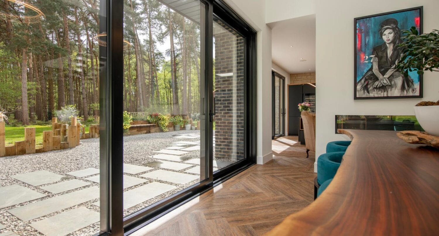 Modern interior with large bespoke glass solutions, featuring sliding doors opening to a stone patio and garden. Wooden herringbone floors, a natural wood counter, green chairs, potted plant, and a colorful portrait complete the scene.