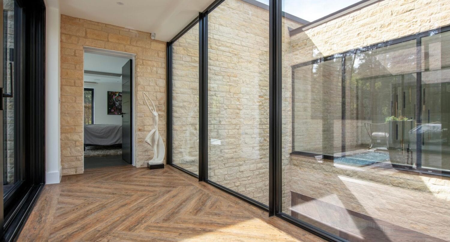 A modern hallway with floor-to-ceiling glass walls, herringbone wood floors, light stone walls, and a view into another room with a bed and artwork visible through open bifold doors.