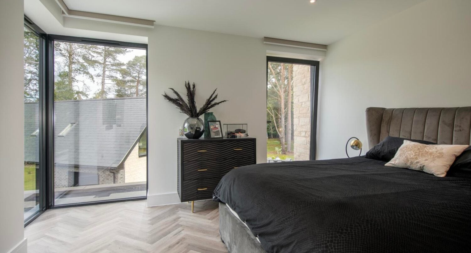 Modern bedroom with large corner windows featuring bespoke glass solutions, black bedspread on a double bed, dark dresser with decorative items, light wood herringbone floor, and a view of trees and neighboring house outside.