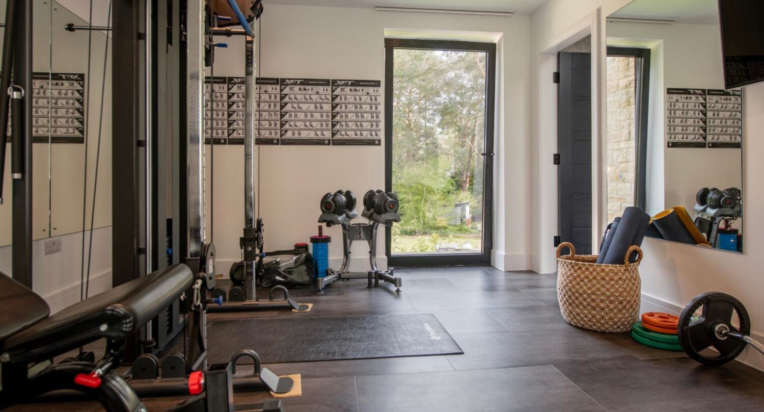 Home gym with various exercise equipment, yoga mats in a basket, and exercise charts on the wall, featuring large windows and bifold doors that let in natural light for an open, inviting space.