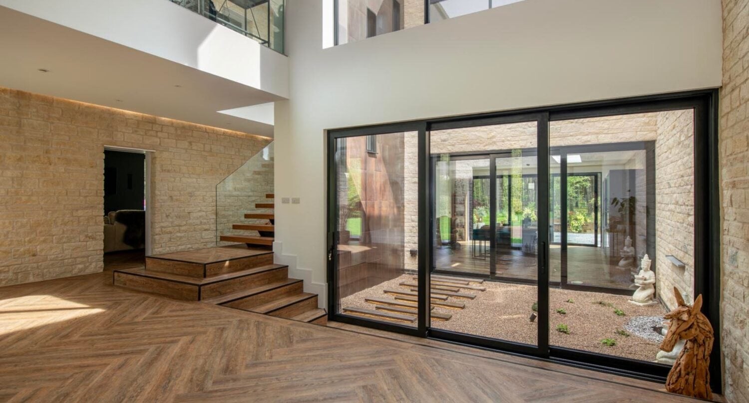 A modern interior features large windows, bespoke glass solutions for the courtyard with gravel and statues, wood herringbone floors, and a floating staircase by stone walls. Natural light fills the open, airy space.