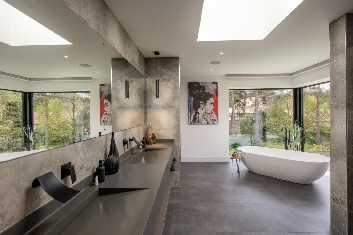Modern bathroom with large mirrors, a double sink, and a freestanding white bathtub by floor-to-ceiling windows and sliding doors, featuring contemporary decor, artwork on the wall, a skylight, and views of greenery outside.