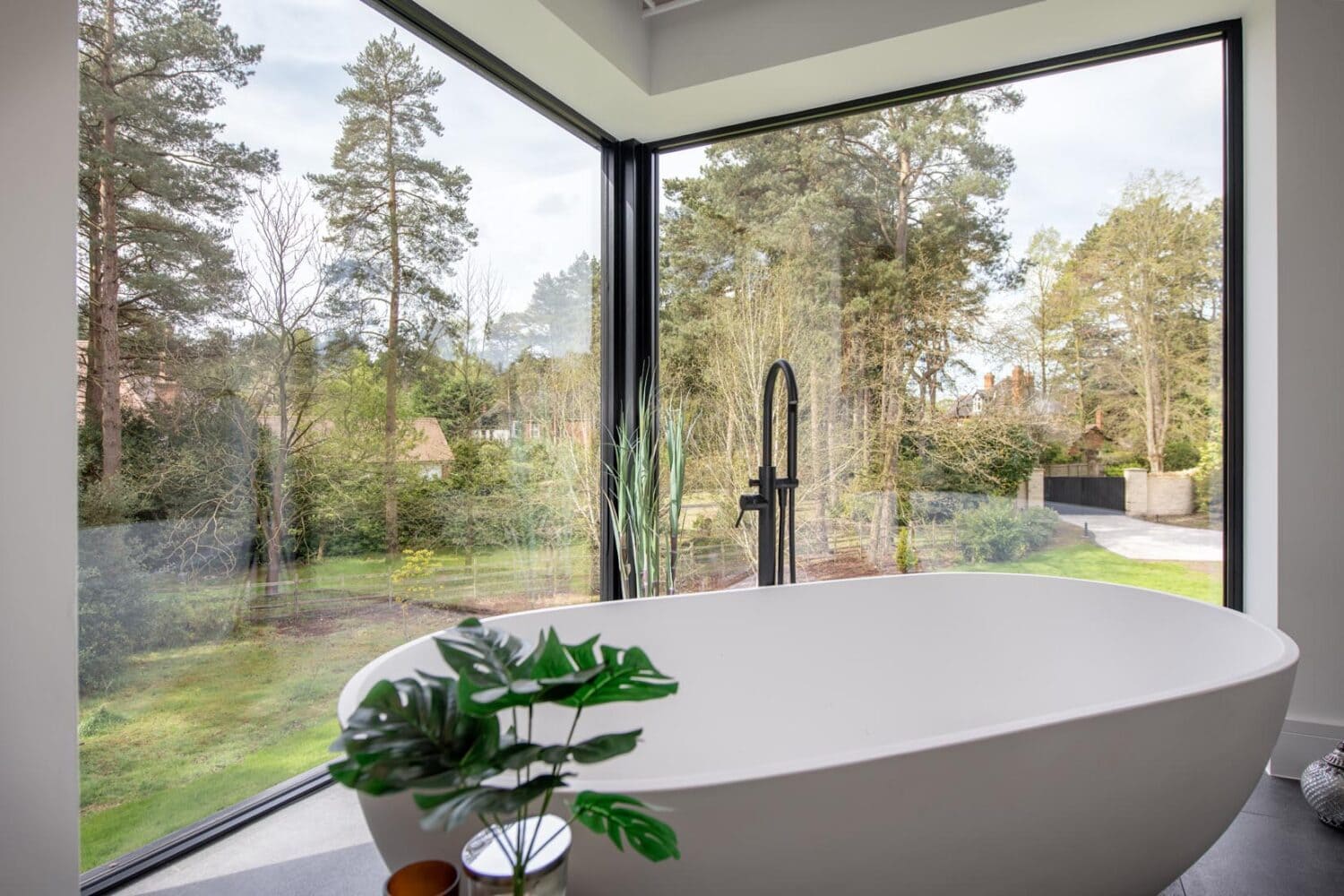 A modern white bathtub is positioned by large floor-to-ceiling corner windows featuring bespoke glass solutions, overlooking a lush green forest. A black faucet stands beside the tub, and a leafy plant decorates the foreground.
