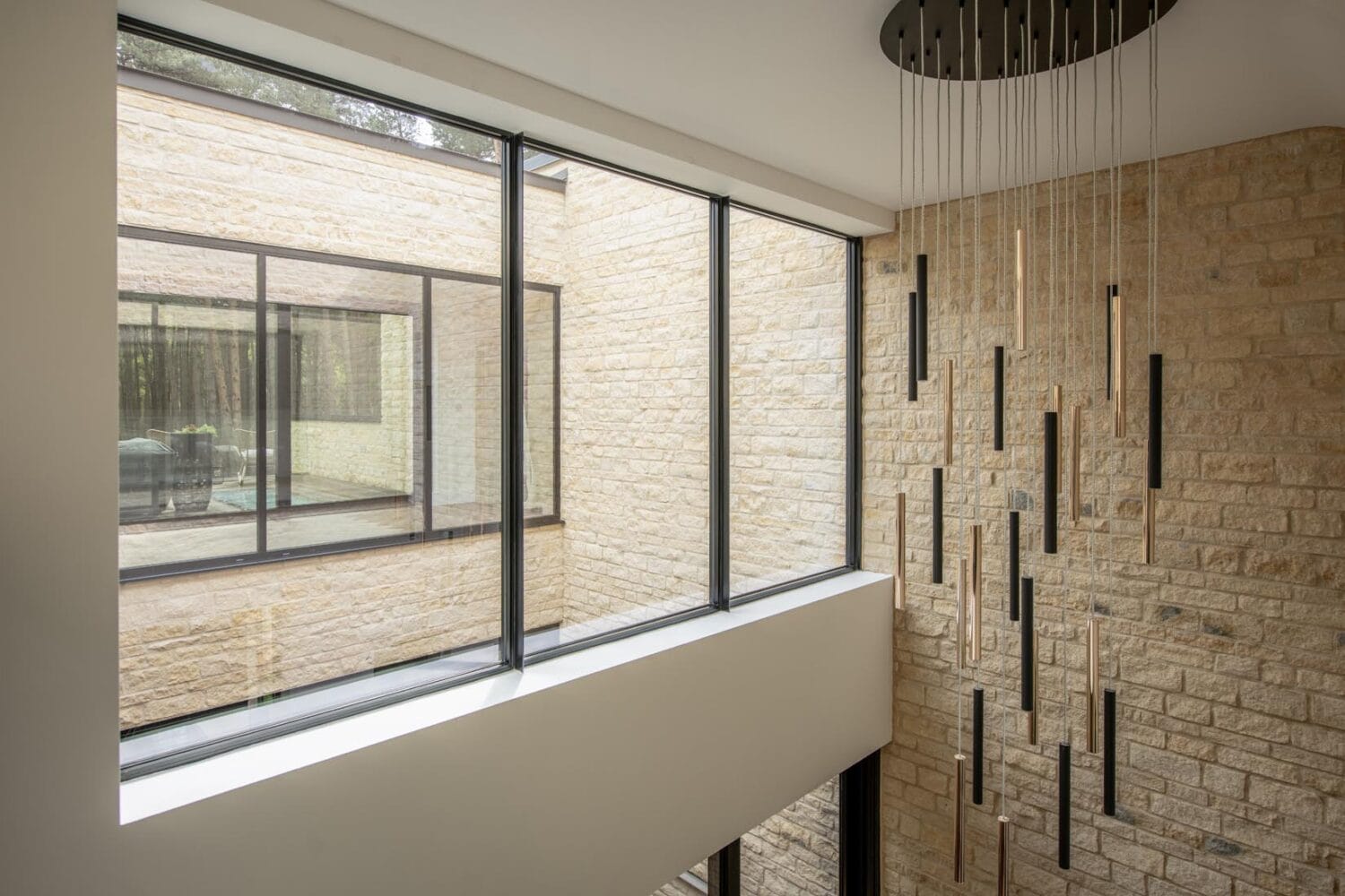 A modern interior with large floor-to-ceiling windows, beige stone walls, and bespoke glass solutions—like contemporary vertical hanging light fixtures—allowing natural light to fill the space.