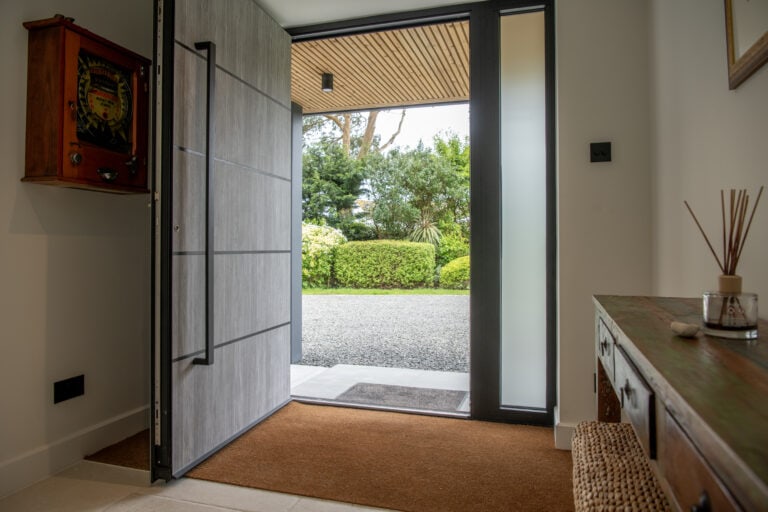 A modern entryway with sliding doors reveals a garden view of green bushes and trees. Inside, there’s a brown mat, a wooden console table, and a reed diffuser.