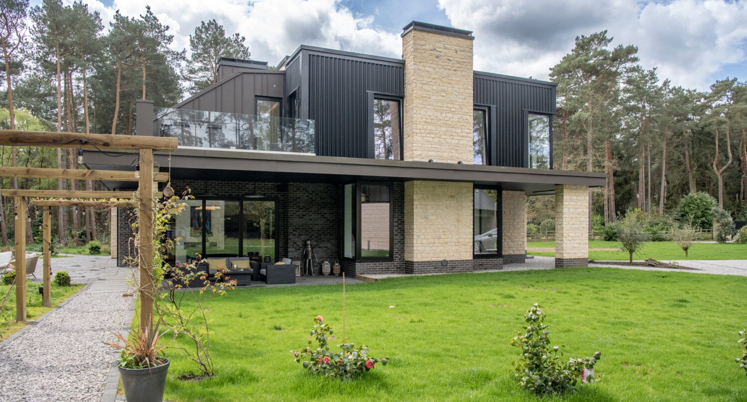 Modern two-story house with black and beige exterior, large windows, sliding doors, a balcony, and a spacious green lawn, surrounded by trees and a garden under a partly cloudy sky.