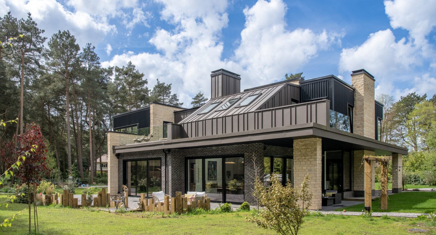 Modern two-story house with large windows featuring bespoke glass solutions, beige brick and dark accents, surrounded by green lawn and trees under a blue sky with clouds. Outdoor seating area is visible near the house.