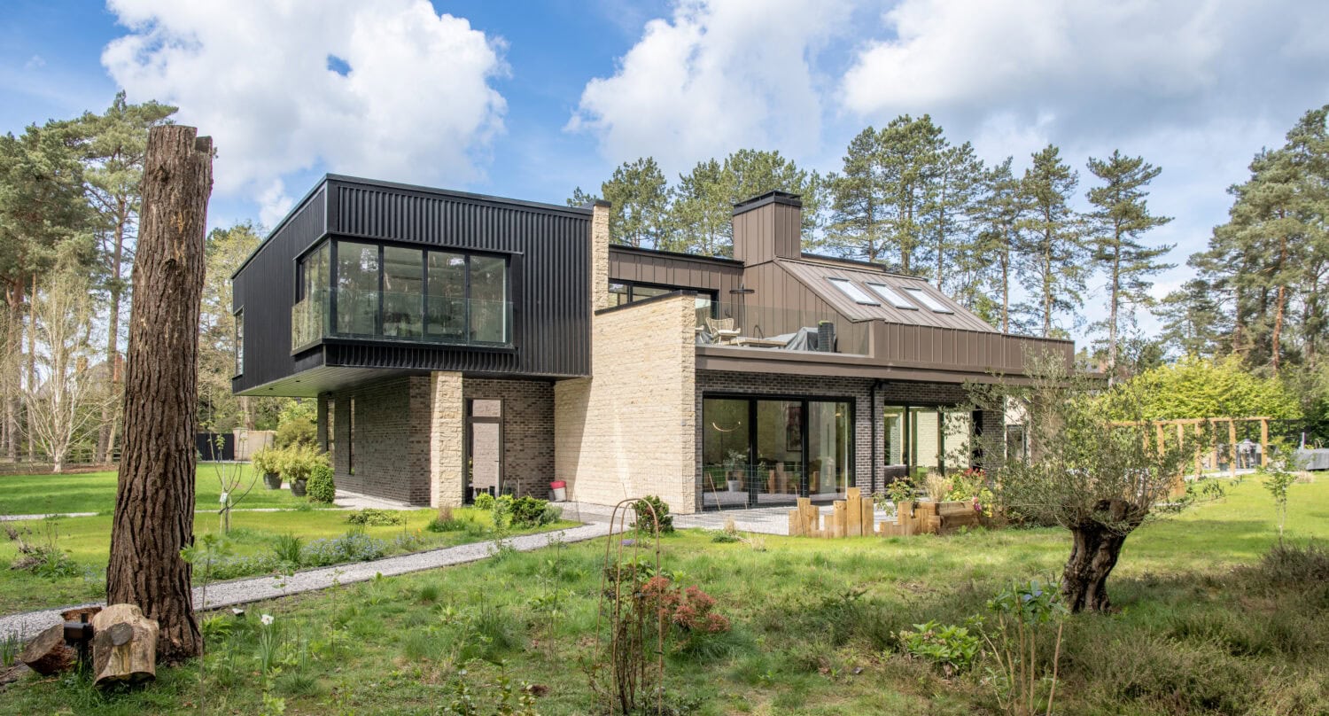 Modern two-story house with large windows, bifold doors, a mix of brick and dark panel siding, and an outdoor patio set in a green, landscaped yard surrounded by tall trees under a partly cloudy sky.