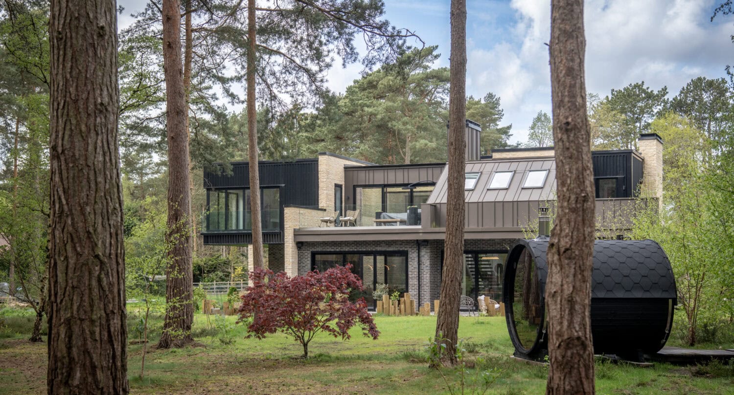 Modern two-story house with large windows featuring bespoke glass solutions and balconies, surrounded by tall trees and greenery. A small red-leafed tree is in the yard, near a round black outdoor structure. The setting is peaceful and wooded.