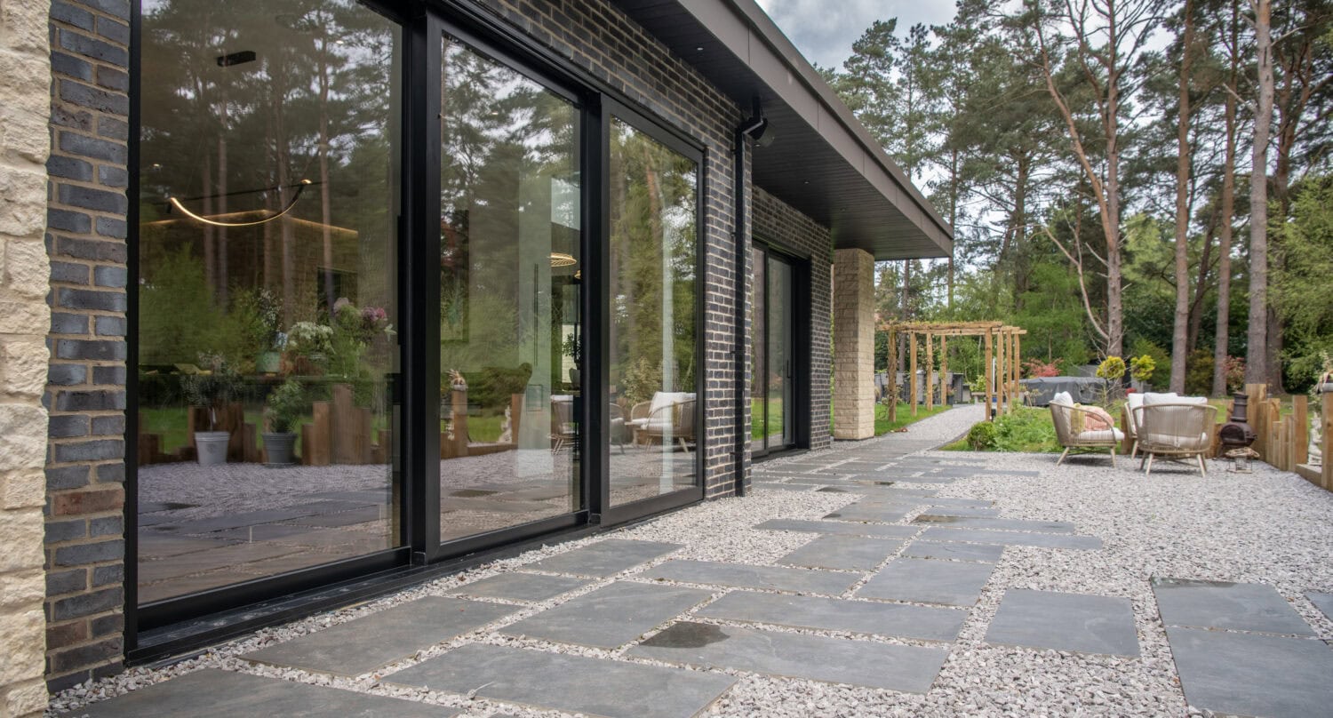 A modern house with bespoke glass solutions features large sliding doors opening onto a patio with square stone tiles, gravel, outdoor seating, plants, and a view of trees and a wooden pergola in the background.