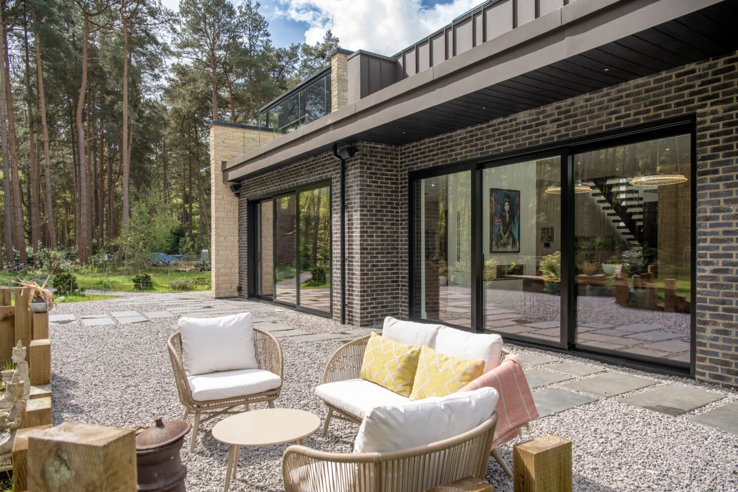 A modern house with dark brick exterior and large bifold doors opens to a patio with outdoor wicker furniture, surrounded by trees and a gravel yard.