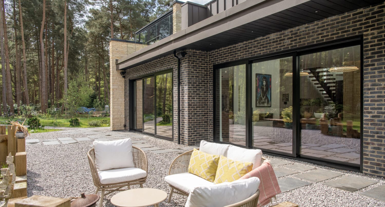 A modern house with dark brick exterior and large bifold doors opens to a patio with outdoor wicker furniture, surrounded by trees and a gravel yard.