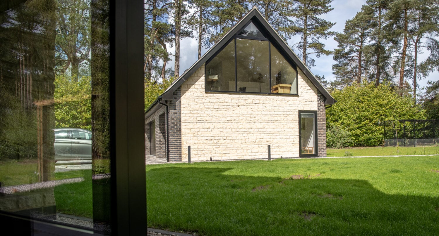 A modern house with light stone walls and large triangular windows sits on a green lawn, surrounded by trees and shrubbery, viewed from inside another building through sleek sliding doors.
