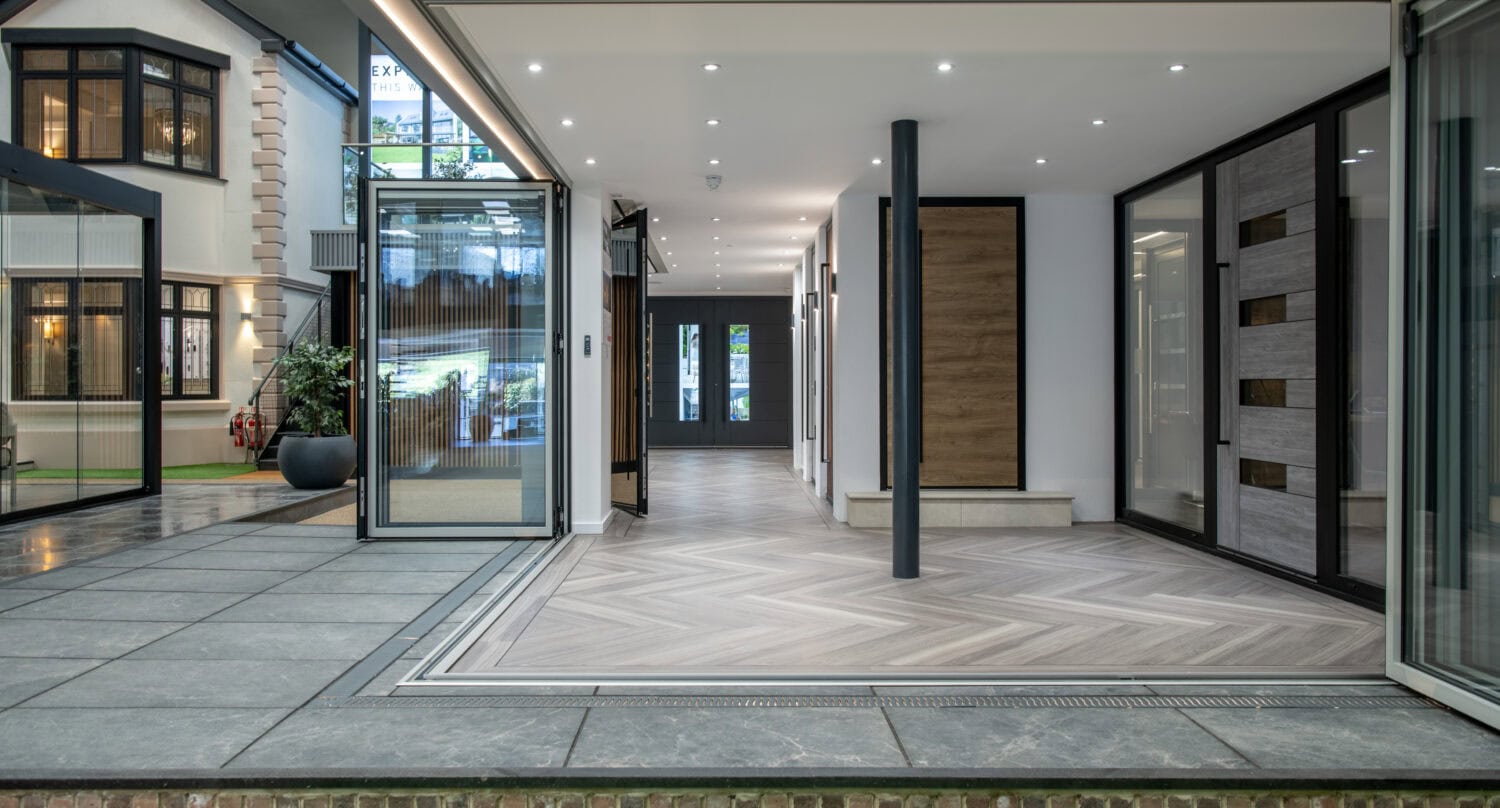 A modern indoor showroom displays contemporary sliding doors, large windows, and wood accents, with tiled flooring and artificial green grass bordering the walkway. Glass roofing brings in natural light for an inviting atmosphere.