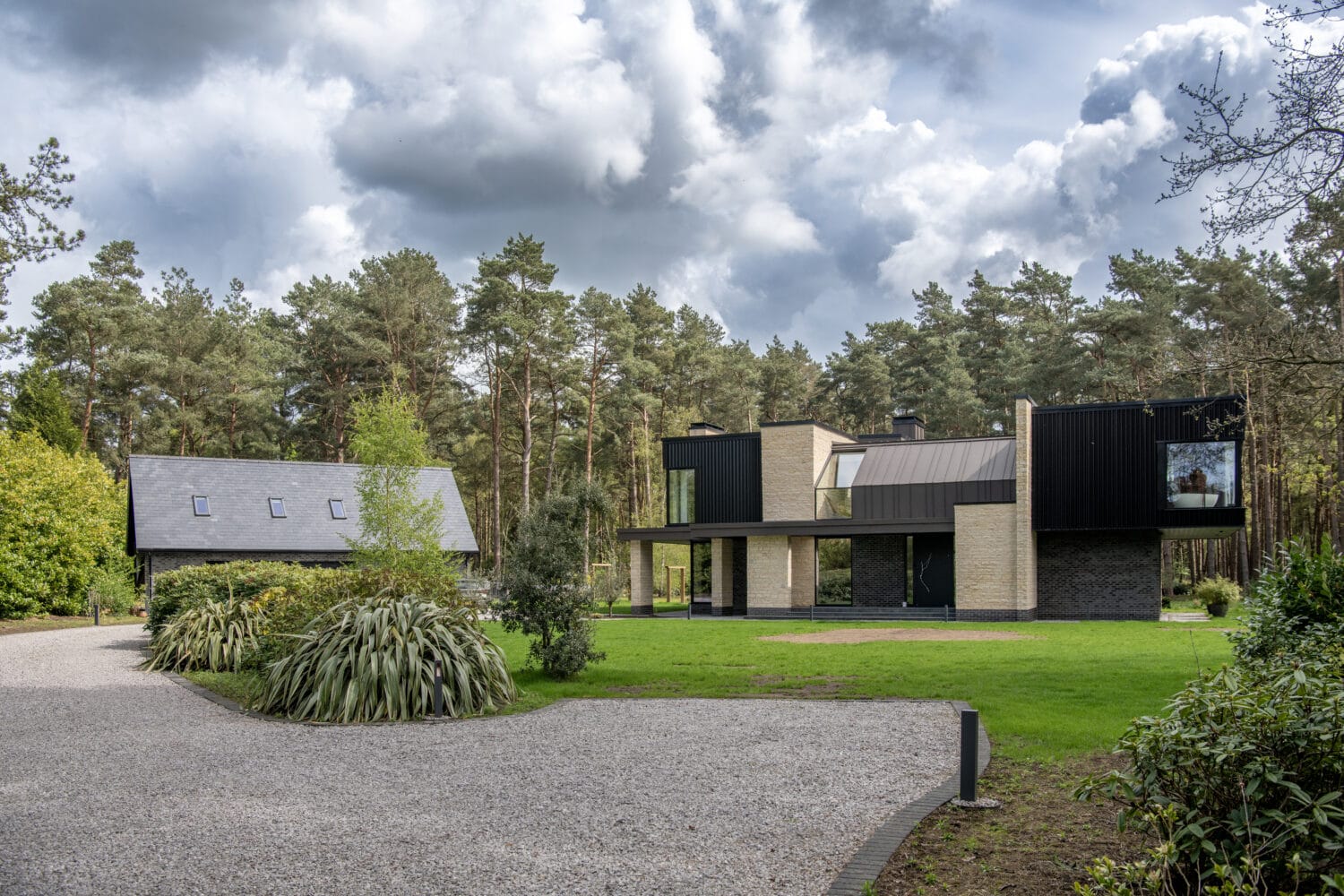 Modern house with bespoke glass solutions, large windows, and a black-and-beige facade sits beside a smaller building with a sloped roof, surrounded by green lawn, trees, and a gravel driveway under a cloudy sky.