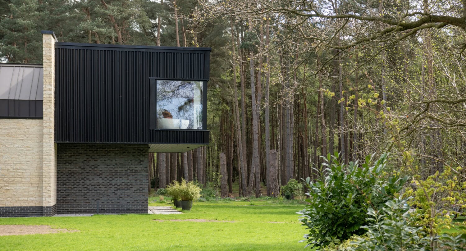 Modern house with black and beige exterior, large window, bifold doors, and a lawn surrounded by green bushes and tall trees in a forest setting.