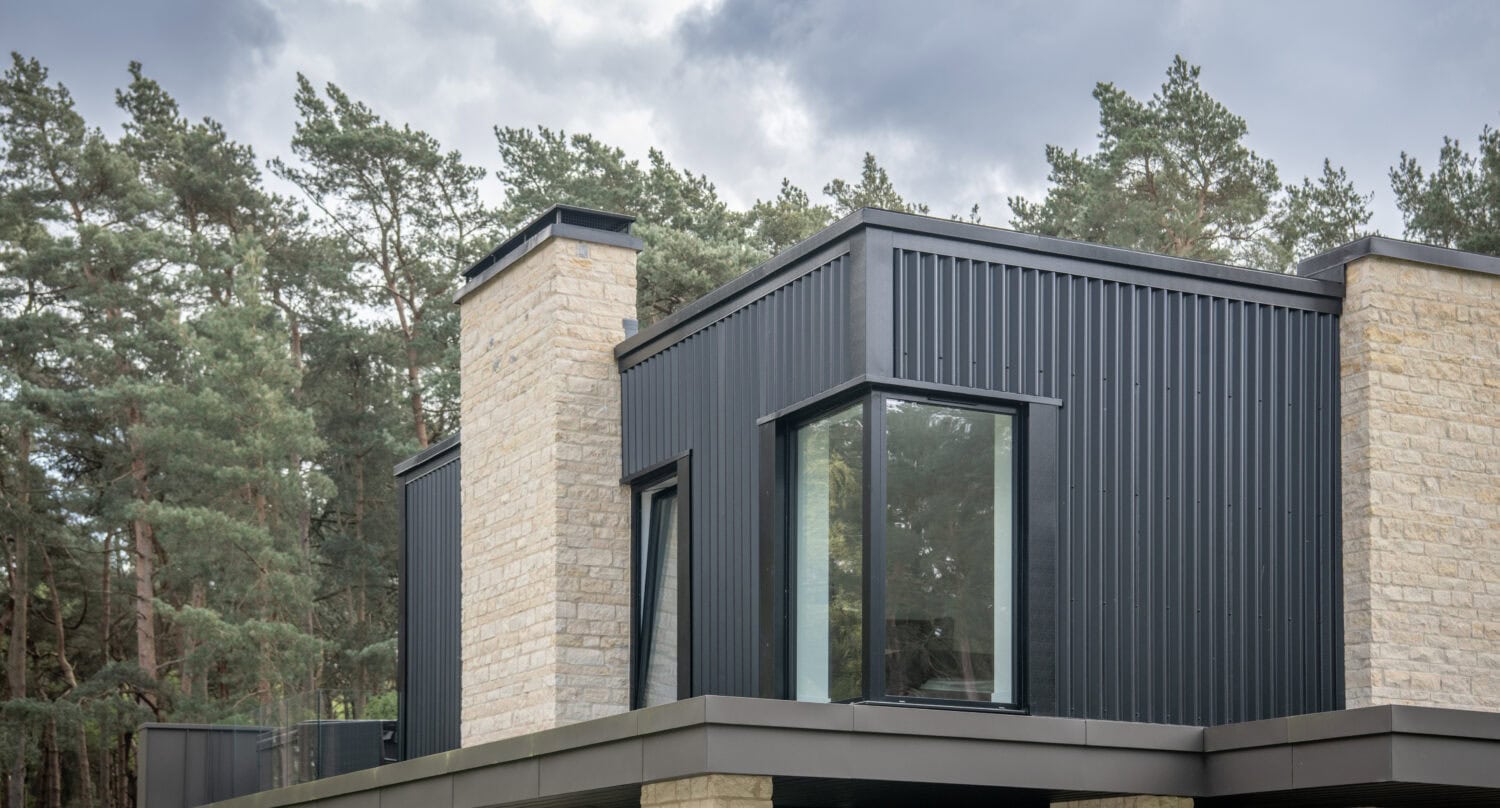 Modern house exterior with large windows, dark vertical metal cladding, light stone walls, and bifold doors, surrounded by pine trees under a cloudy sky. The building has a boxy, minimalist design.