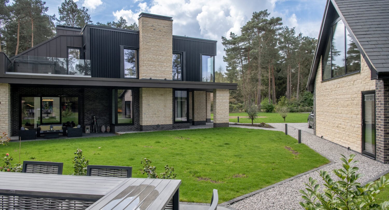 Modern two-story house with stone and dark accents, large windows, sliding doors, a balcony, and a detached garage. A patio with an outdoor table and green lawn are in the foreground, surrounded by trees and blue sky with clouds.