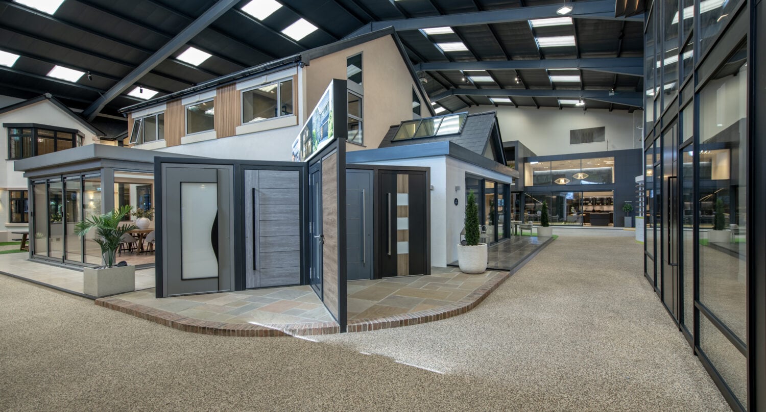 Modern indoor showroom featuring full-scale house models and a variety of exterior and bifold doors on display, with ample lighting from overhead skylights and large windows, plus clean walkways throughout the space.