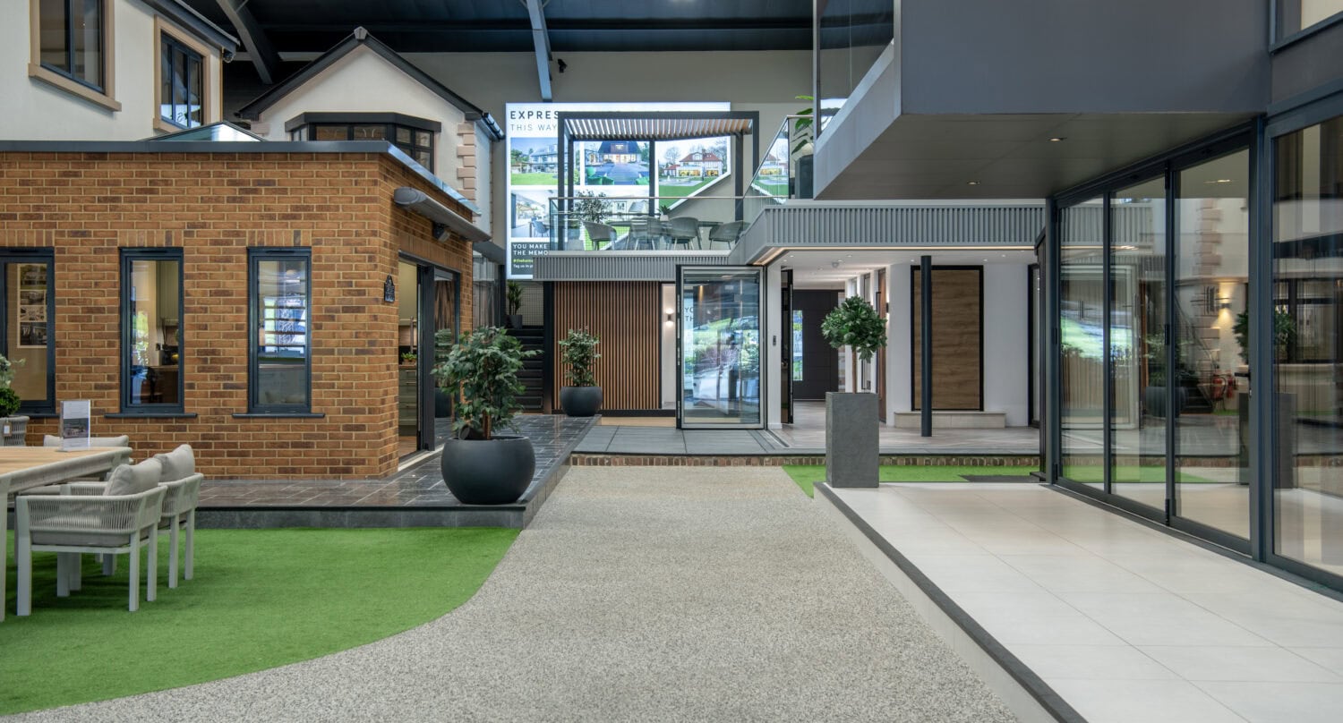 A modern indoor display area features model homes, large windows, bifold doors, outdoor furniture, potted plants, and bright lighting, showcasing various architectural and landscaping styles.