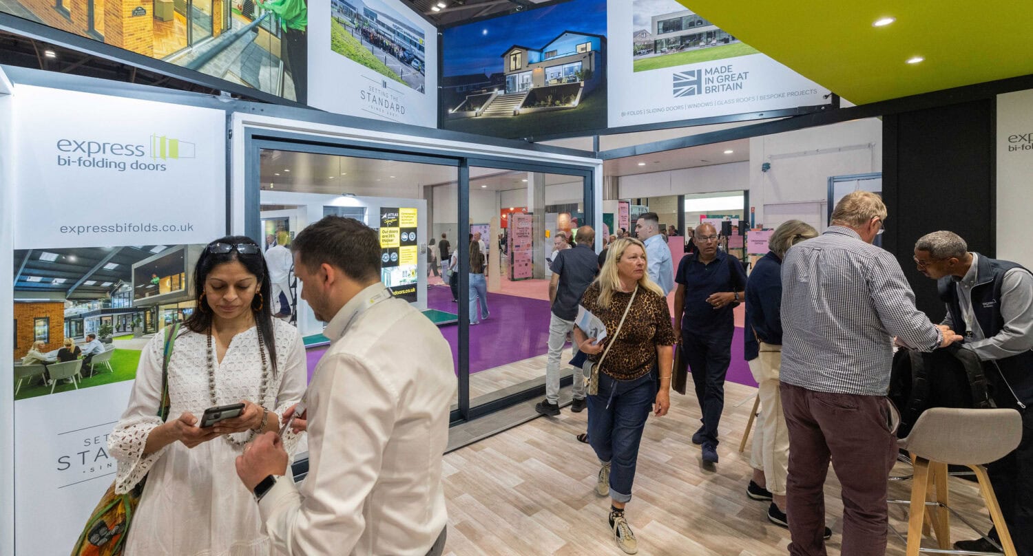 People visit an exhibition booth for Express Bi-Folding Doors. Some talk and look at brochures on bifold doors, while others walk past. Large displays show photos of modern homes with front doors and glass roofing in the background.