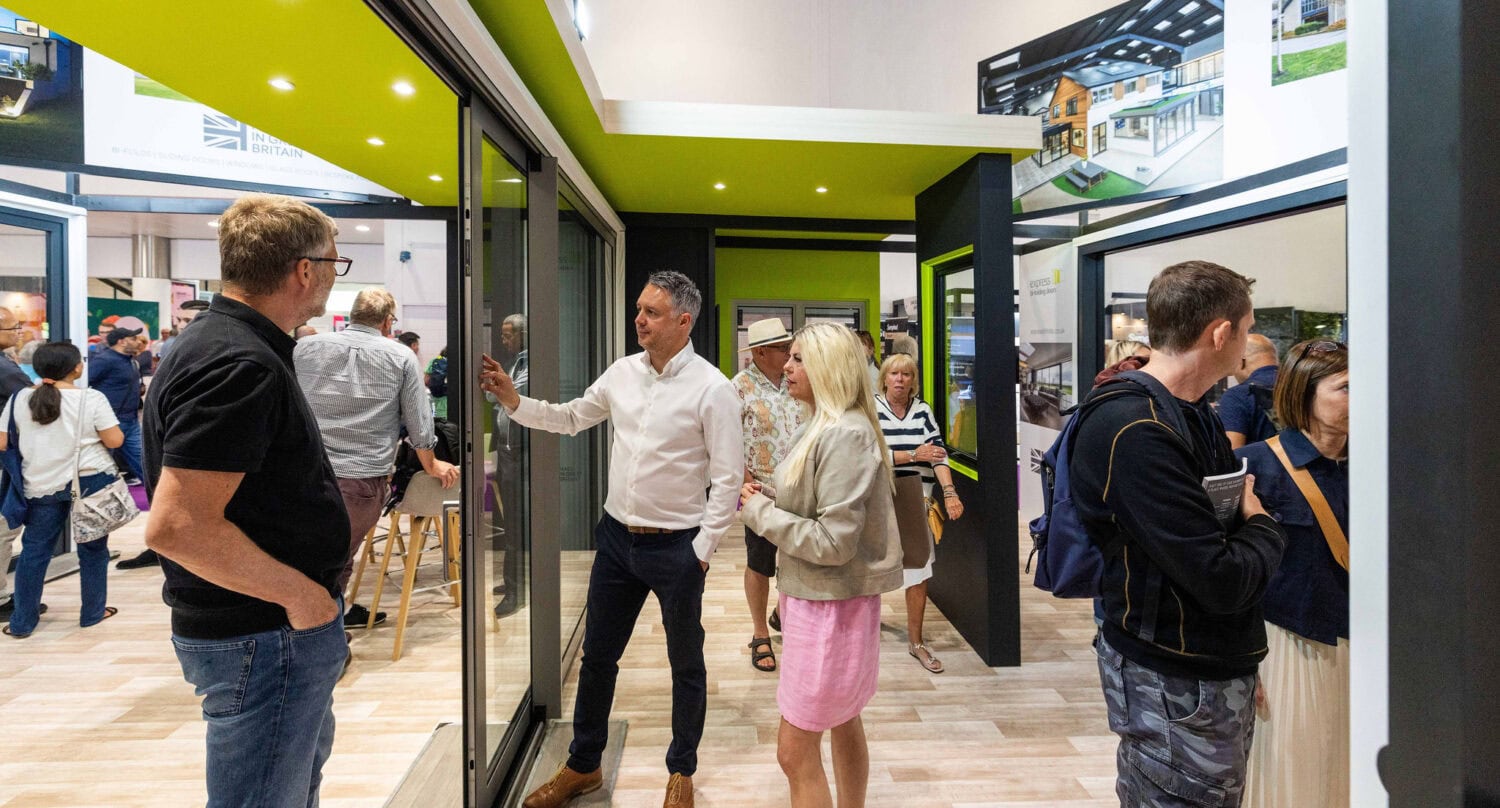 People interact and examine features in a modern showroom with bespoke glass solutions, including large glass doors and green accents. Some stand and talk while others look around; informational displays and photos are visible in the background.