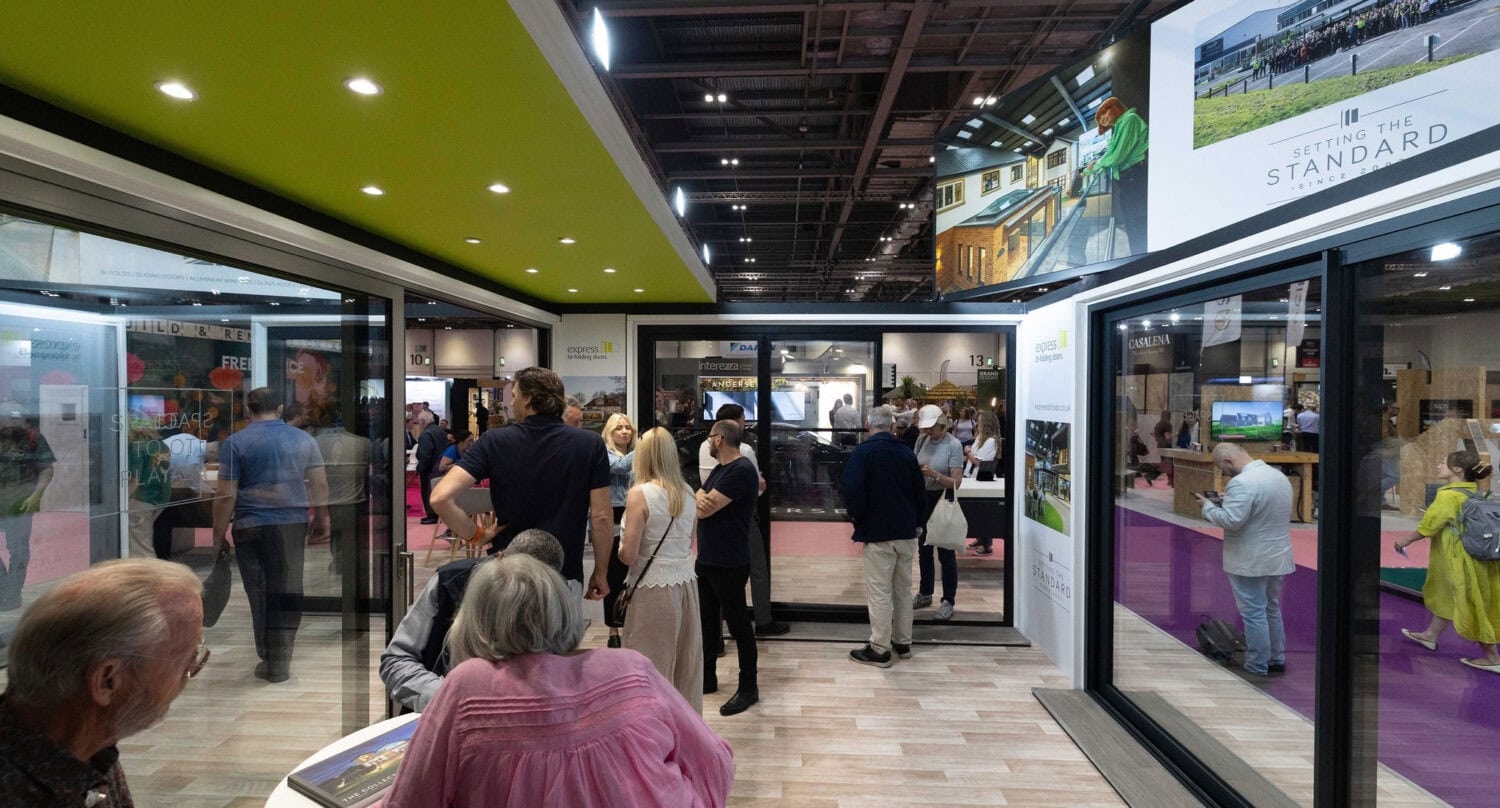 People visit a modern exhibition hall with bespoke glass solutions, featuring glass walls and informational displays. Some are seated at tables, while others walk around and view exhibits. The atmosphere is lively and spacious.