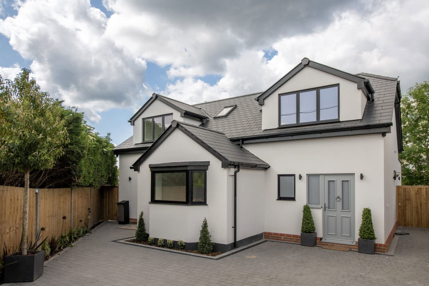 Modern two-story house with white walls, grey roof, and large windows. There is a paved driveway, a few small shrubs by the entrance, wooden fencing, and trees along the side, with a cloudy sky overhead.
