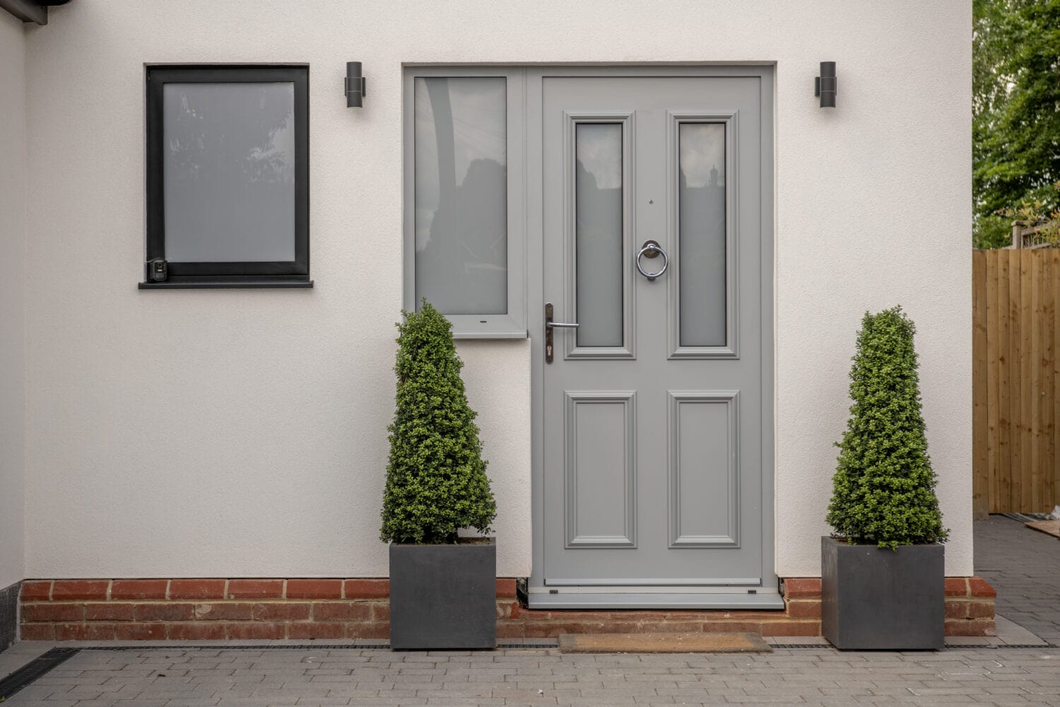 A modern front door painted light gray with geometric panels, flanked by two tall, cone-shaped potted shrubs. The house exterior is white with a black-framed window and minimalist wall lights.
