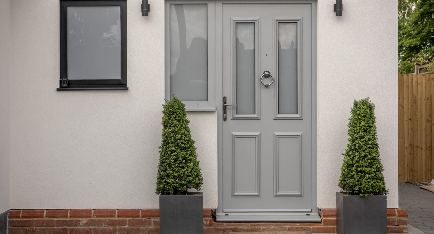 A modern front door painted light gray with geometric panels, flanked by two tall, cone-shaped potted shrubs. The house exterior is white with a black-framed window and minimalist wall lights.