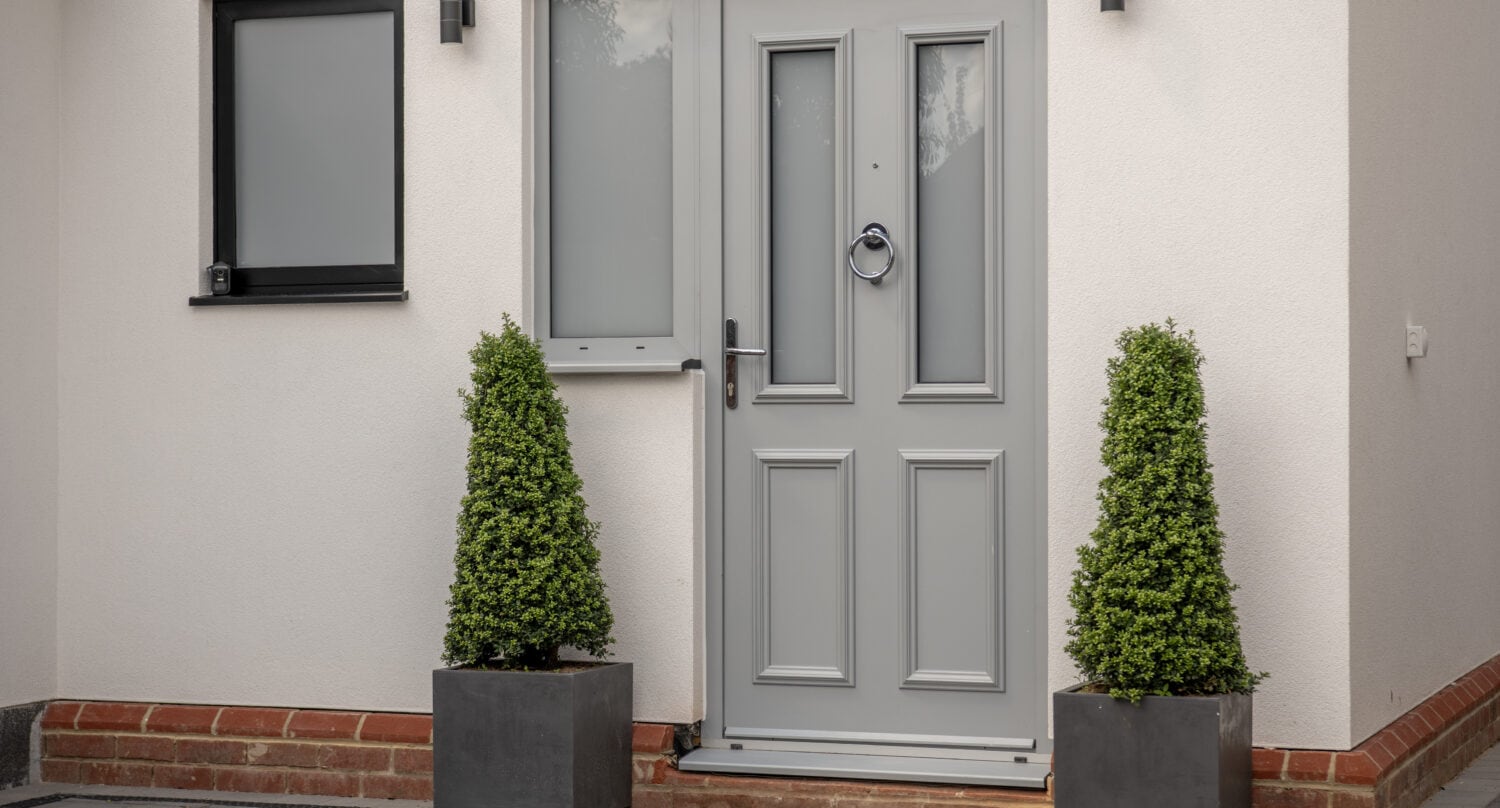 A modern light gray front door with frosted glass panels, flanked by two tall, cone-shaped evergreen shrubs in square planters. The door is set in a white exterior wall with a small window to the left.