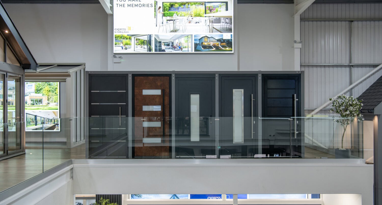 A modern showroom interior with stylish bifold doors displayed on an upper level behind glass railings. Above the doors, a lighted sign reads YOU MAKE THE MEMORIES with home interior photos and bespoke glass solutions throughout the space.