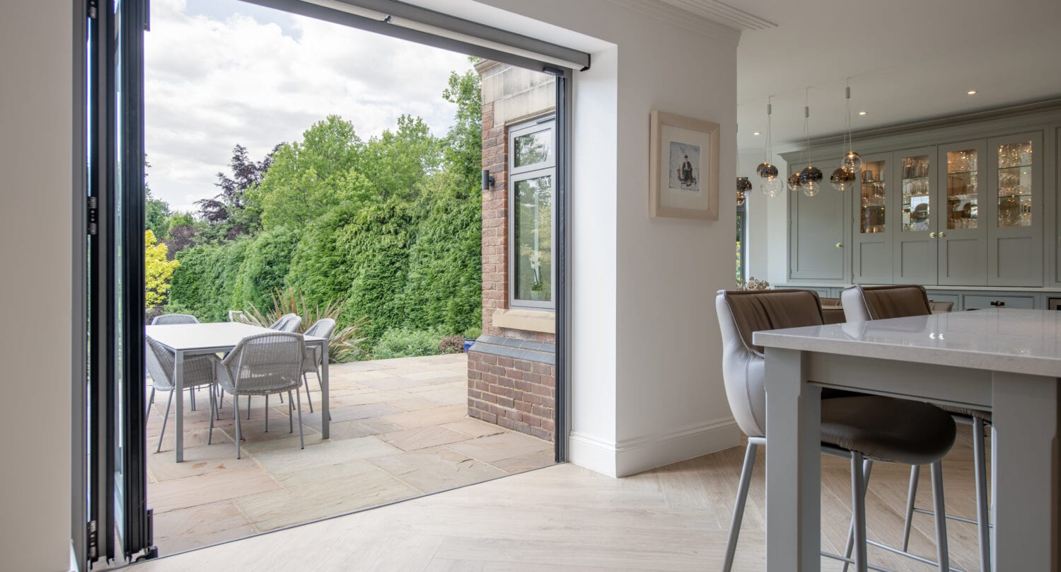 Modern kitchen with an island and bar stools opens to a patio through large folding glass doors. Outdoor dining table and chairs are set on a paved terrace, overlooking a lush green garden.